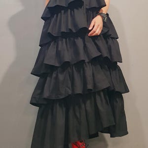 May include: A black sleeveless dress with a high collar and a zipper closure. The dress has multiple layers of ruffles and a long, flowing skirt. The model is wearing red lace-up platform sandals.
