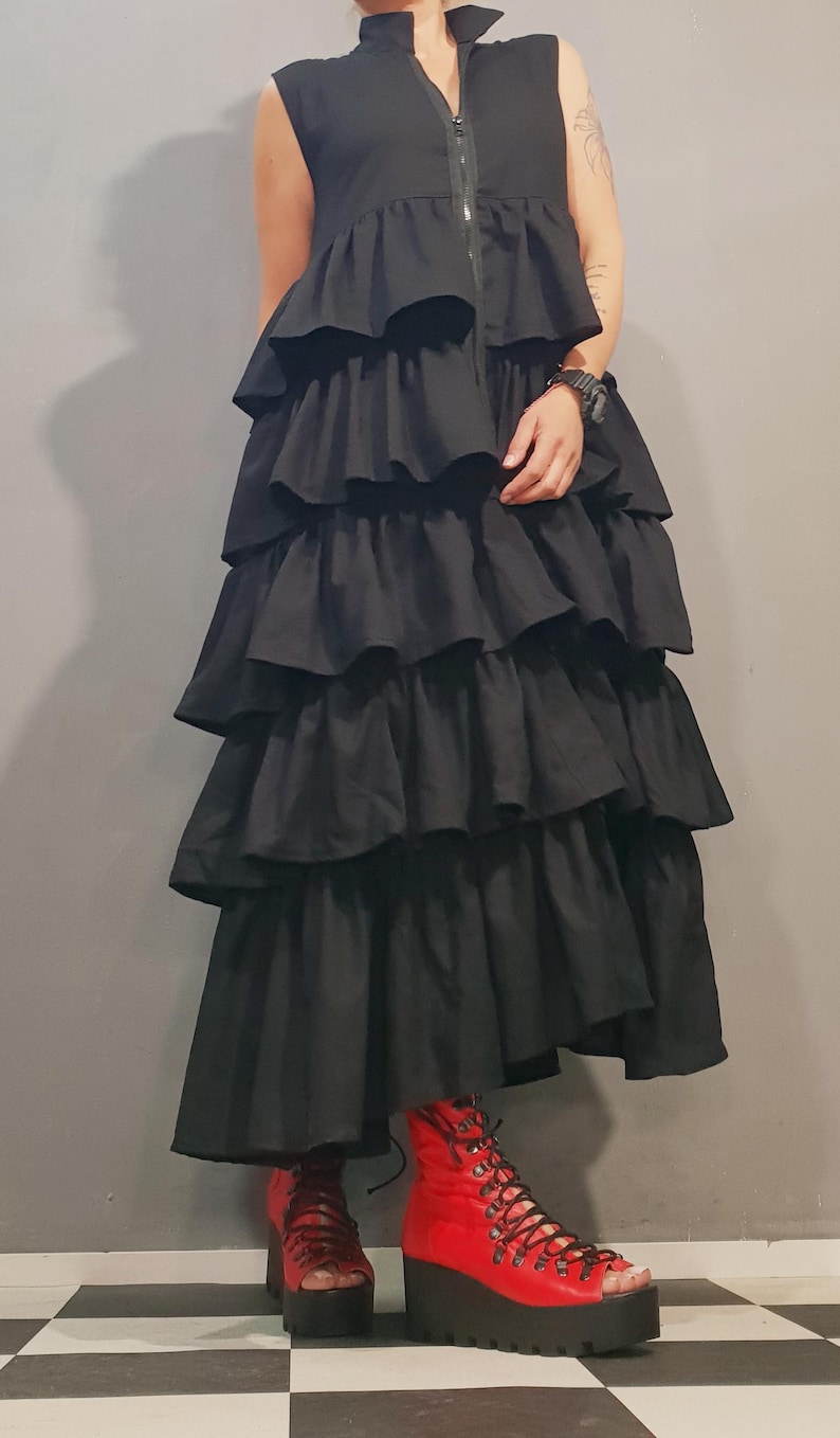 May include: A black sleeveless dress with a high collar and a zipper closure. The dress has multiple layers of ruffles and a long, flowing skirt. The model is wearing red lace-up platform sandals.