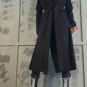 May include: A long black coat with a wide collar, a belt, and two large pockets. The coat is worn over black pants and white platform boots.