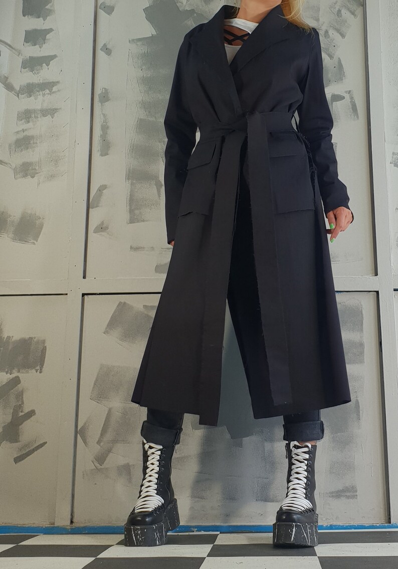 May include: A long black coat with a wide collar, a belt, and two large pockets. The coat is worn over black pants and white platform boots.