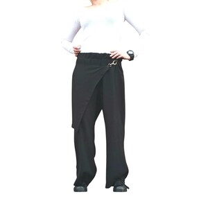 May include: Black wrap-style pants with a unique asymmetrical design. The pants feature a high waist and a decorative gold-colored clasp. The fabric appears to be a lightweight material, suitable for warmer weather. The pants are paired with a white long-sleeve top.