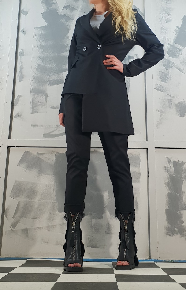 May include: A black blazer with a double-breasted closure and a long, asymmetrical hemline. The blazer is paired with black pants and black heeled boots with zippers.