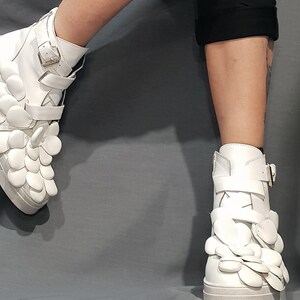 May include: White high-top sneakers with a unique design featuring multiple white flower-shaped embellishments on the sides and a buckle closure.