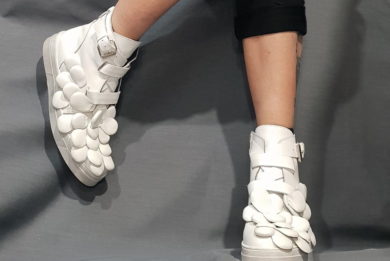 May include: White high-top sneakers with a unique design featuring multiple white flower-shaped embellishments on the sides and a buckle closure.