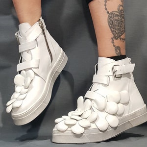May include: White high-top sneakers with a unique design featuring multiple white flower-shaped embellishments on the front of the shoe.