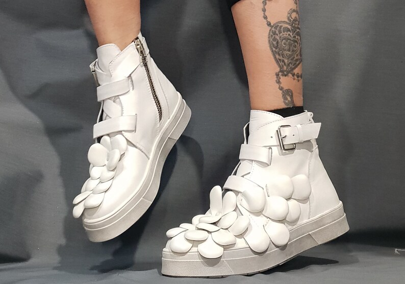May include: White high-top sneakers with a unique design featuring multiple white flower-shaped embellishments on the front of the shoe.