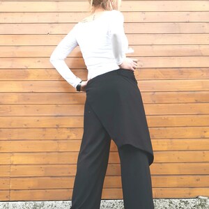 May include: Black wrap-style pants with a wide leg design. The pants are paired with a long-sleeved white top. The model is standing in front of a wooden wall.