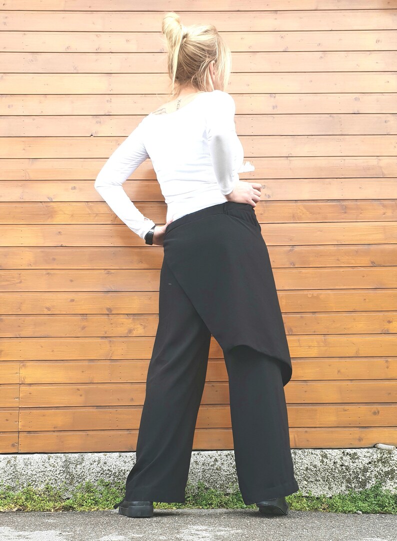 May include: Black wrap-style pants with a wide leg design. The pants are paired with a long-sleeved white top. The model is standing in front of a wooden wall.