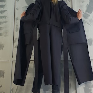 May include: A black long-sleeved jacket with long ties hanging from the waist. The jacket is worn by a person with blonde hair, and the person is standing in front of a white wall with gray paint splatters.