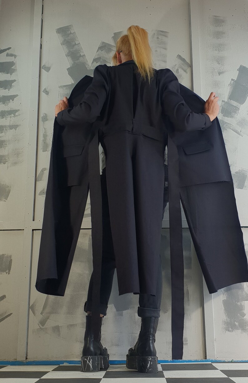 May include: A black long-sleeved jacket with long ties hanging from the waist. The jacket is worn by a person with blonde hair, and the person is standing in front of a white wall with gray paint splatters.