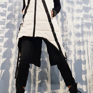May include: A white puffer vest with black diagonal stripes and a black zipper. The vest has a hood and is worn over a black long-sleeved shirt and black pants. The person is wearing black lace-up boots.