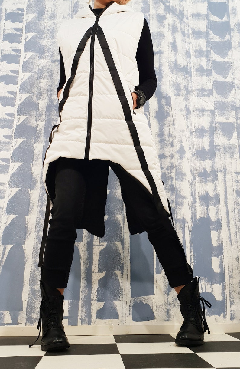 May include: A white puffer vest with black diagonal stripes and a black zipper. The vest has a hood and is worn over a black long-sleeved shirt and black pants. The person is wearing black lace-up boots.