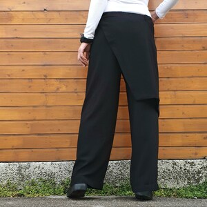 May include: Black wrap-style pants with a wide-leg design. The pants feature a unique overlapping fabric detail at the front. The model is wearing a white long-sleeve shirt and black shoes. The background is a wooden wall.