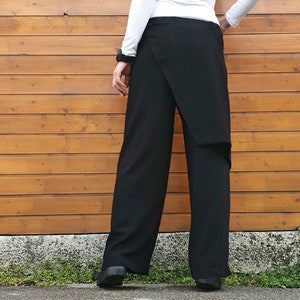 May include: Black wrap-style pants with a wide-leg design. The pants feature a unique overlapping fabric detail at the front. The model is wearing a white long-sleeve shirt and black shoes. The background is a wooden wall.