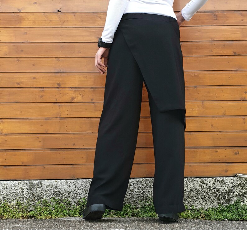 May include: Black wrap-style pants with a wide-leg design. The pants feature a unique overlapping fabric detail at the front. The model is wearing a white long-sleeve shirt and black shoes. The background is a wooden wall.