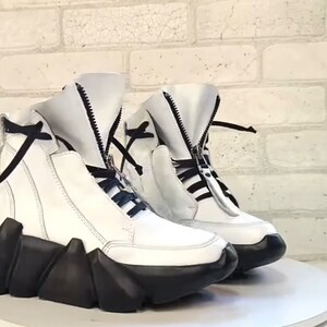 May include: A pair of white leather high-top sneakers with black accents. The sneakers feature a unique design with a black, geometric sole and a zipper closure. Black laces and a black tie detail the top of the shoe.