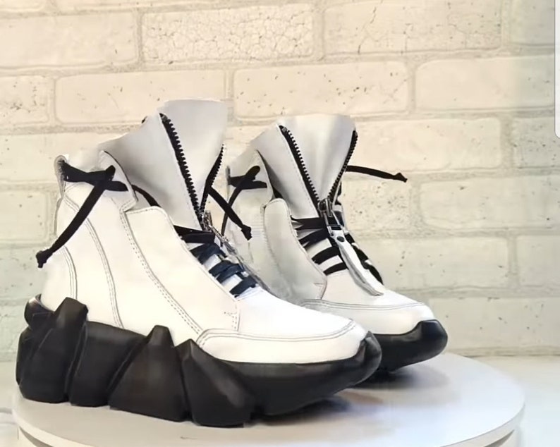 May include: A pair of white leather high-top sneakers with black accents. The sneakers feature a unique design with a black, geometric sole and a zipper closure. Black laces and a black tie detail the top of the shoe.