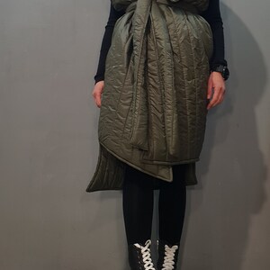 May include: A long, olive green quilted vest with a cinched waist and a high collar. The vest is worn over black leggings and black platform boots with white laces.
