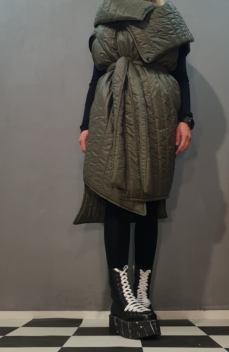 May include: A long, olive green quilted vest with a cinched waist and a high collar. The vest is worn over black leggings and black platform boots with white laces.