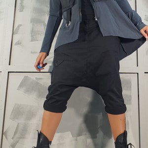 May include: A person wearing a black quilted vest over a gray long-sleeved shirt and black pants. The pants have a unique design with a gathered crotch and cuffed legs. The person is wearing black lace-up boots with thick soles.