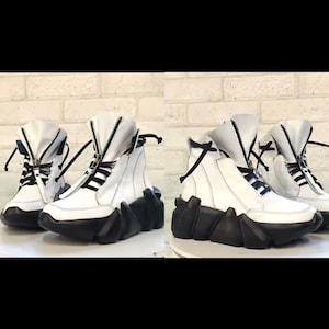 May include: White high-top sneakers with black soles and zippers. The sneakers have a unique, geometric design on the soles and black laces. The uppers are white leather, and the shoes are displayed against a white brick wall.