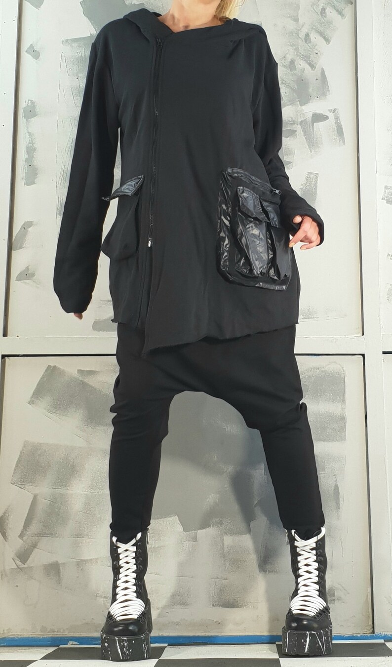 Op de afbeelding: Zwarte hoodie met een rits en een grote zwarte zak aan de voorkant. De jas wordt gedragen over een zwarte broek en zwarte platformlaarzen met witte veters.