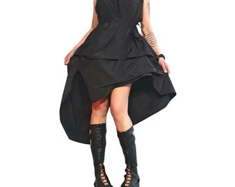 May include: A sleeveless black dress featuring a high-low hemline with layered fabric panels and a tie-waist detail. The dress is paired with black knee-high platform boots that have an open-toe design and lace-up detailing along the front.