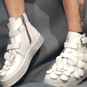 May include: White high-top sneakers with a unique design featuring multiple white flower-shaped embellishments on the front of the shoe.