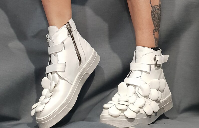 May include: White high-top sneakers with a unique design featuring multiple white flower-shaped embellishments on the front of the shoe.