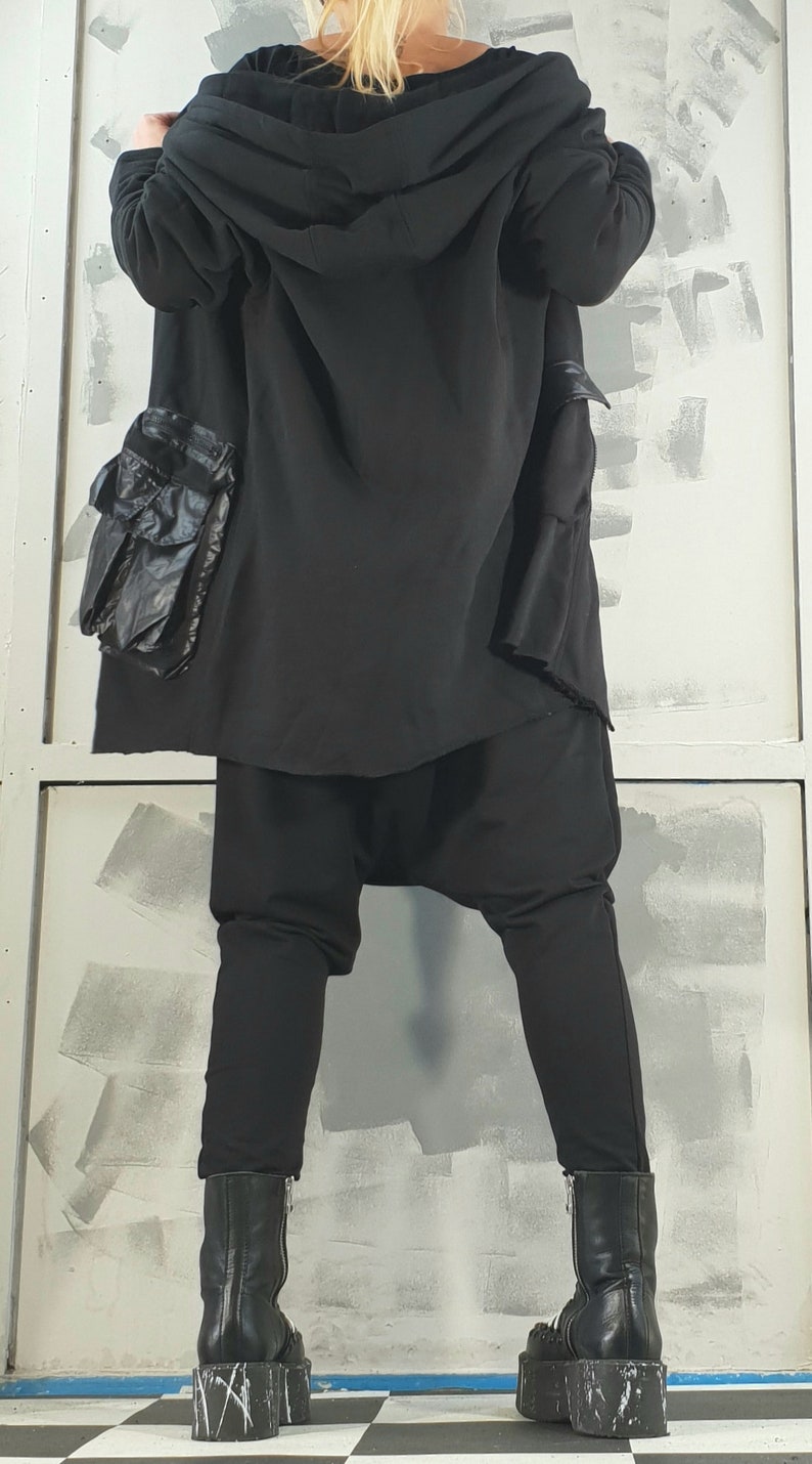 Op de afbeelding: Een zwarte hoodie met een lang, vloeiend ontwerp. De jas heeft een grote zak aan de linkerkant en wordt gedragen met een zwarte broek en zwarte platformlaarzen.