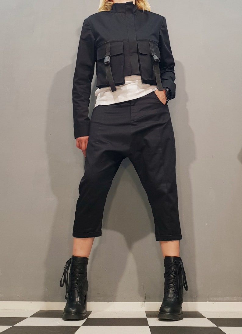 May include: A black cropped jacket with a high collar and multiple straps and pockets. The jacket is paired with black cropped pants and black lace-up boots.
