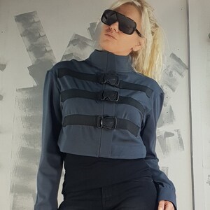 May include: A dark blue cropped jacket with black straps and buckles. The jacket has a high collar and long sleeves.