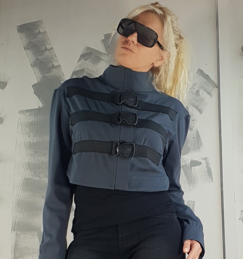 May include: A dark blue cropped jacket with black straps and buckles. The jacket has a high collar and long sleeves.