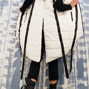 May include: A white puffer vest with black straps and a hood. The vest is long and has a loose fit. The straps are tied at the waist and the vest is open in the front.