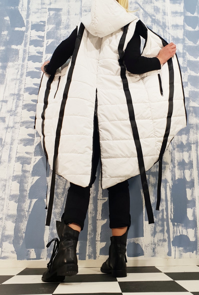 May include: A white puffer vest with black straps and a hood. The vest is long and has a loose fit. The straps are tied at the waist and the vest is open in the front.