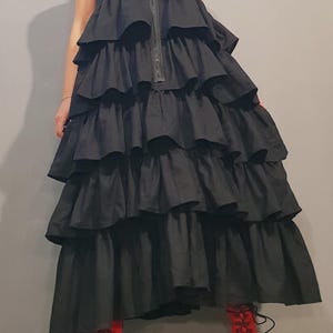 May include: A black sleeveless dress with a high neckline and a zipper down the front. The dress has multiple layers of ruffles and a long, flowing skirt. The model is wearing red platform boots.