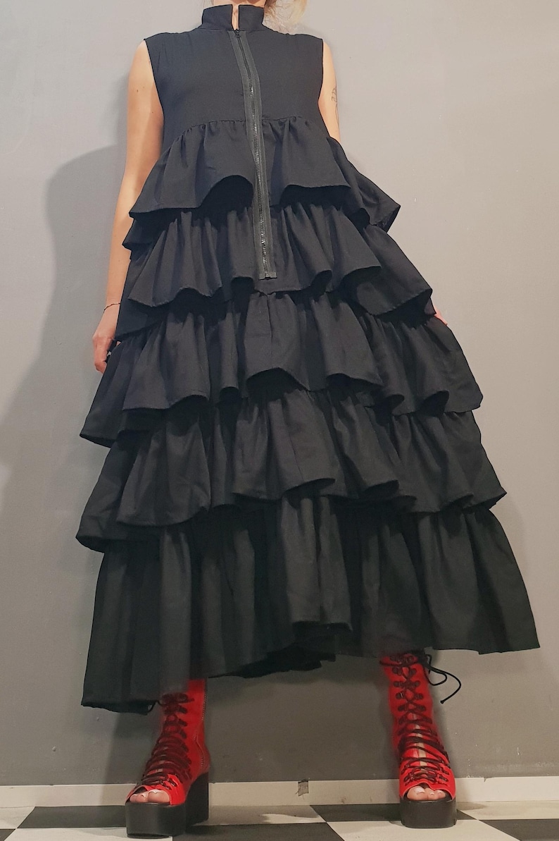 May include: A black sleeveless dress with a high neckline and a zipper down the front. The dress has multiple layers of ruffles and a long, flowing skirt. The model is wearing red platform boots.