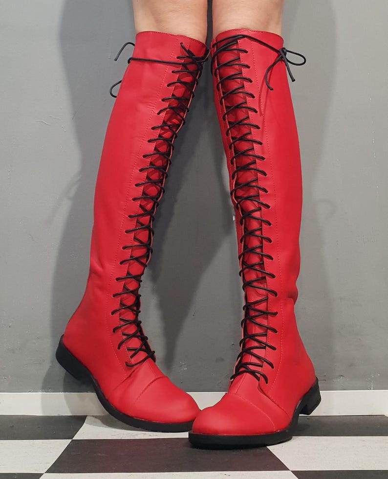 May include: A pair of red knee-high boots with black laces. The boots have a flat sole and a smooth, leather-like texture. The laces run up the front of the boots, creating a striking visual contrast.