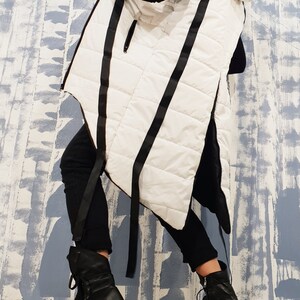 May include: A white hooded vest with black straps and a quilted design. The vest is long and has a asymmetrical hemline. The model is wearing black pants and black boots.