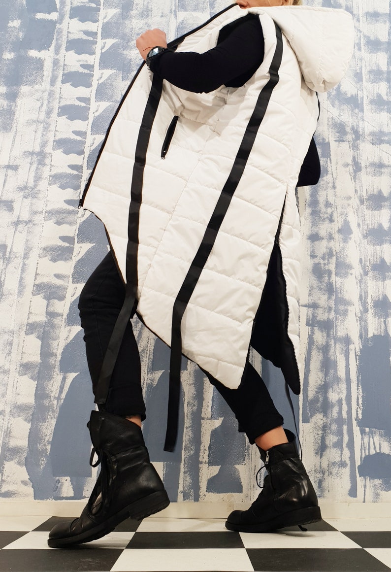 May include: A white hooded vest with black straps and a quilted design. The vest is long and has a asymmetrical hemline. The model is wearing black pants and black boots.