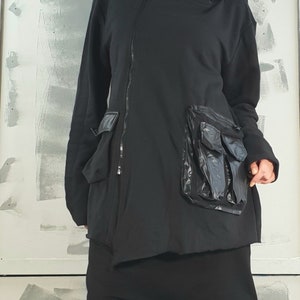 Op de afbeelding: Zwarte hoodie met een lange rits en een groot, doorzichtig plastic zakje aan de voorkant.