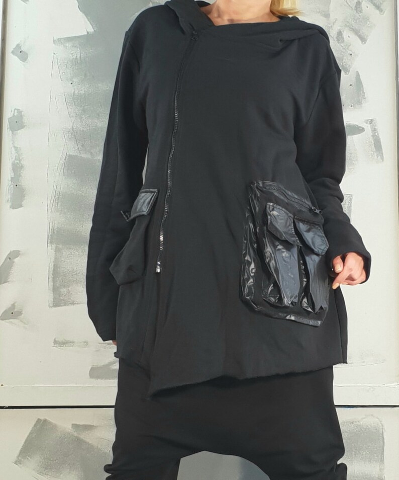Op de afbeelding: Zwarte hoodie met een lange rits en een groot, doorzichtig plastic zakje aan de voorkant.
