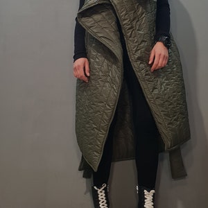May include: A long, olive green quilted vest with a large, folded collar. The vest has a cinched waist and is worn over black trousers and black platform boots with white laces.