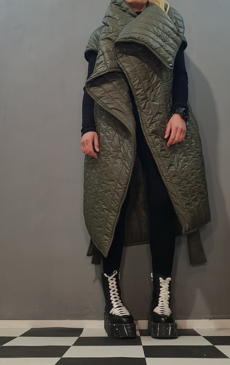 May include: A long, olive green quilted vest with a large, folded collar. The vest has a cinched waist and is worn over black trousers and black platform boots with white laces.