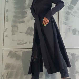 May include: A black long-sleeved coat with a belt and a long slit on each side. The coat is worn over black jeans and black and white platform boots.