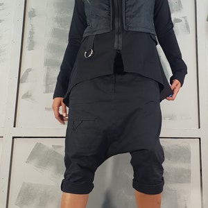 May include: A woman wearing a black hooded vest, black pants, and black boots. The vest has a zipper and a silver ring on the front. The pants are loose-fitting and have a zipper on the side. The boots are lace-up and have a thick platform.