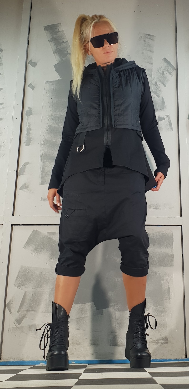 May include: A woman wearing a black hooded vest, black pants, and black boots. The vest has a zipper and a silver ring on the front. The pants are loose-fitting and have a zipper on the side. The boots are lace-up and have a thick platform.