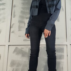May include: A woman wearing a blue cropped jacket with black buckles, black jeans, and black heeled boots. She is wearing sunglasses and has her hair pulled back.