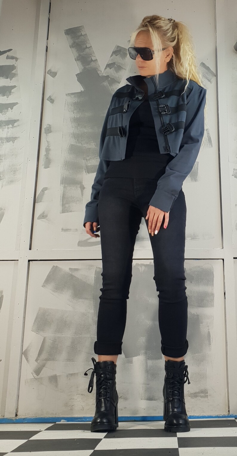 May include: A woman wearing a blue cropped jacket with black buckles, black jeans, and black heeled boots. She is wearing sunglasses and has her hair pulled back.