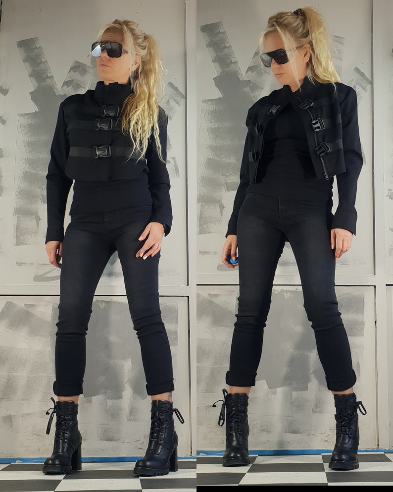 May include: A black cropped jacket with multiple buckles and a black turtleneck shirt. The model is wearing black jeans and black heeled boots.